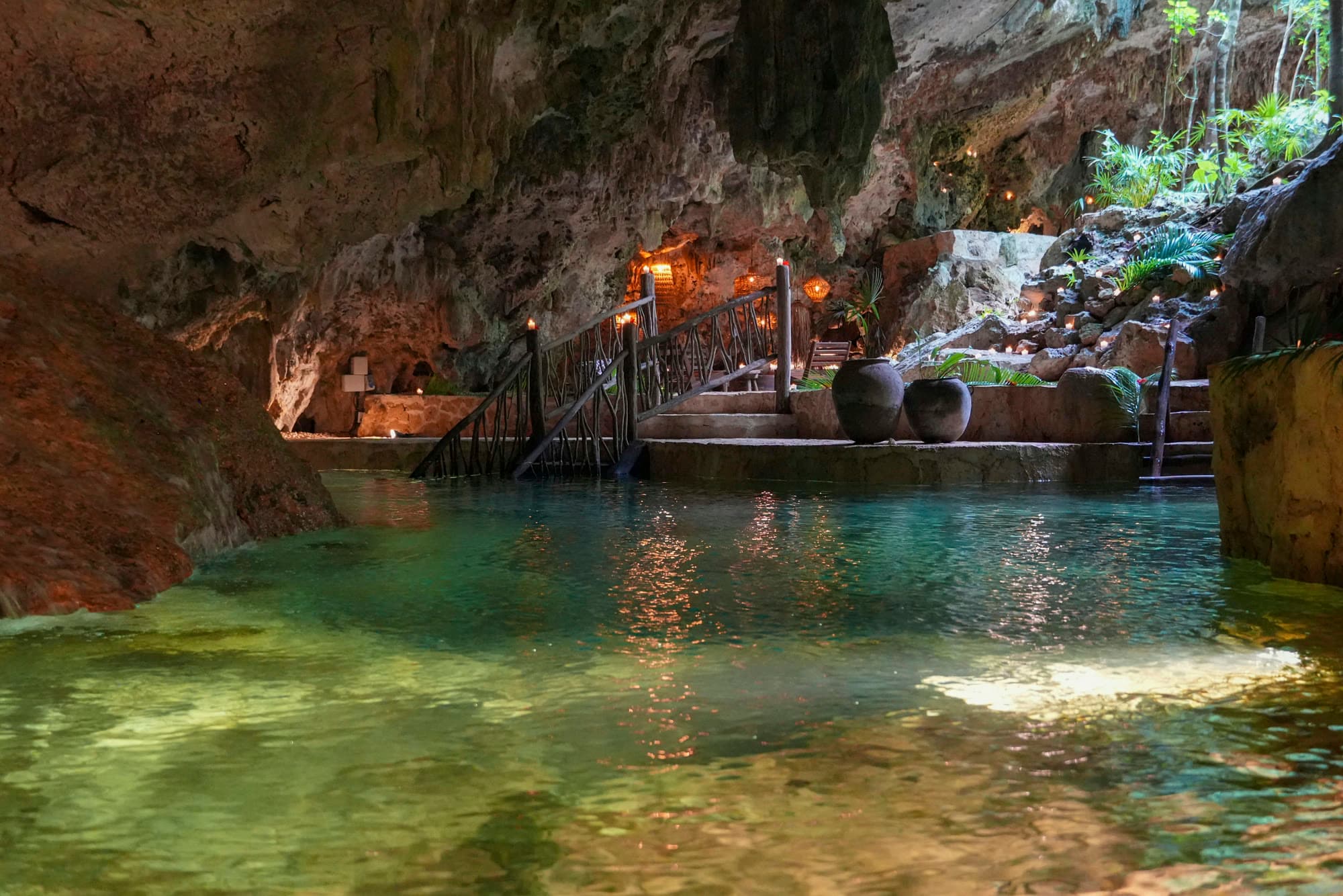 The cenote by candlelight: the turquoise pool, the lanterns along the rock and the wooden walkway at the water's edge