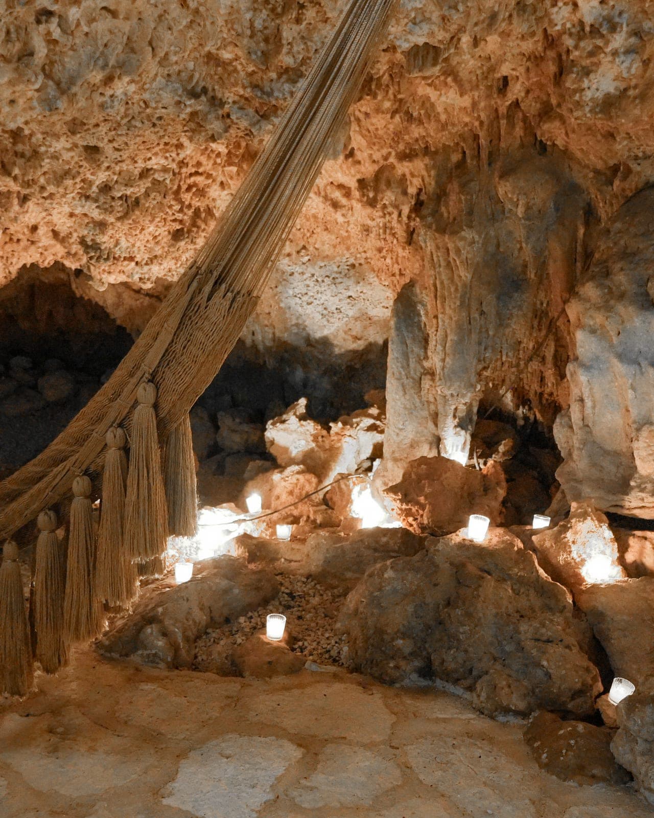 A hammock strung between stalagmites in the candlelit cave, votive flames scattered across the rock