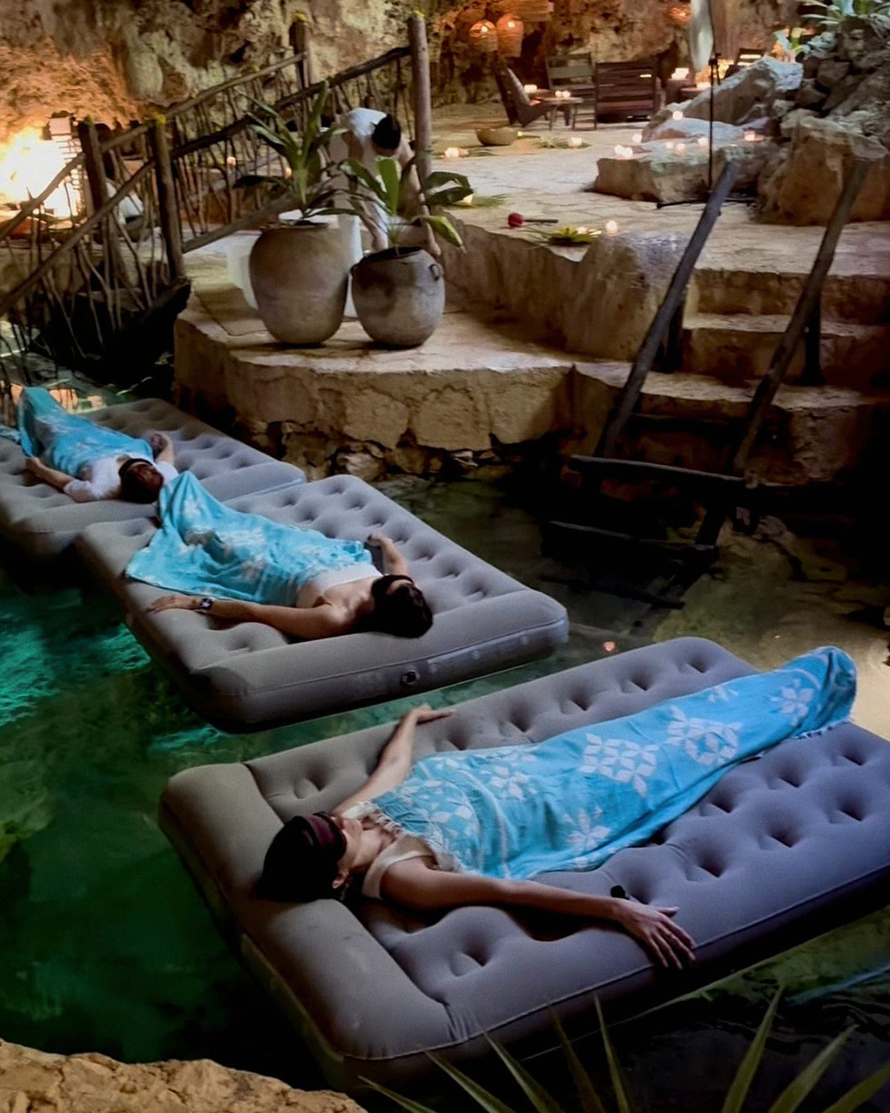 Guests wrapped in blankets floating on cushioned mattresses across the cenote, an attendant tending candles by the bridge