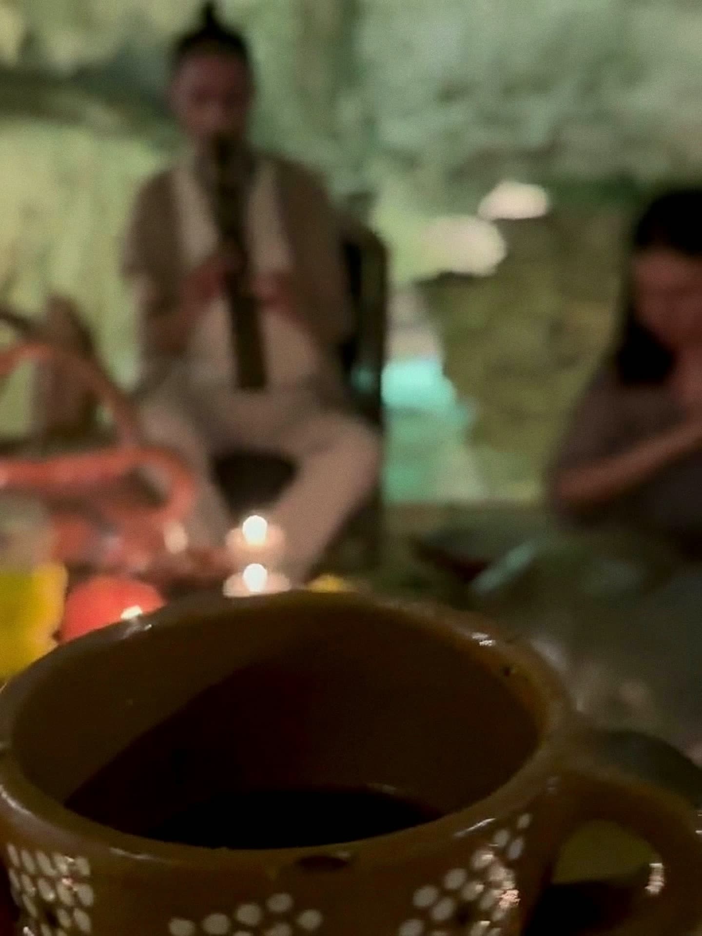 A Medicine Musician playing a wooden pan flute on a stool in the cave, candles and offerings at his feet and the cenote behind him