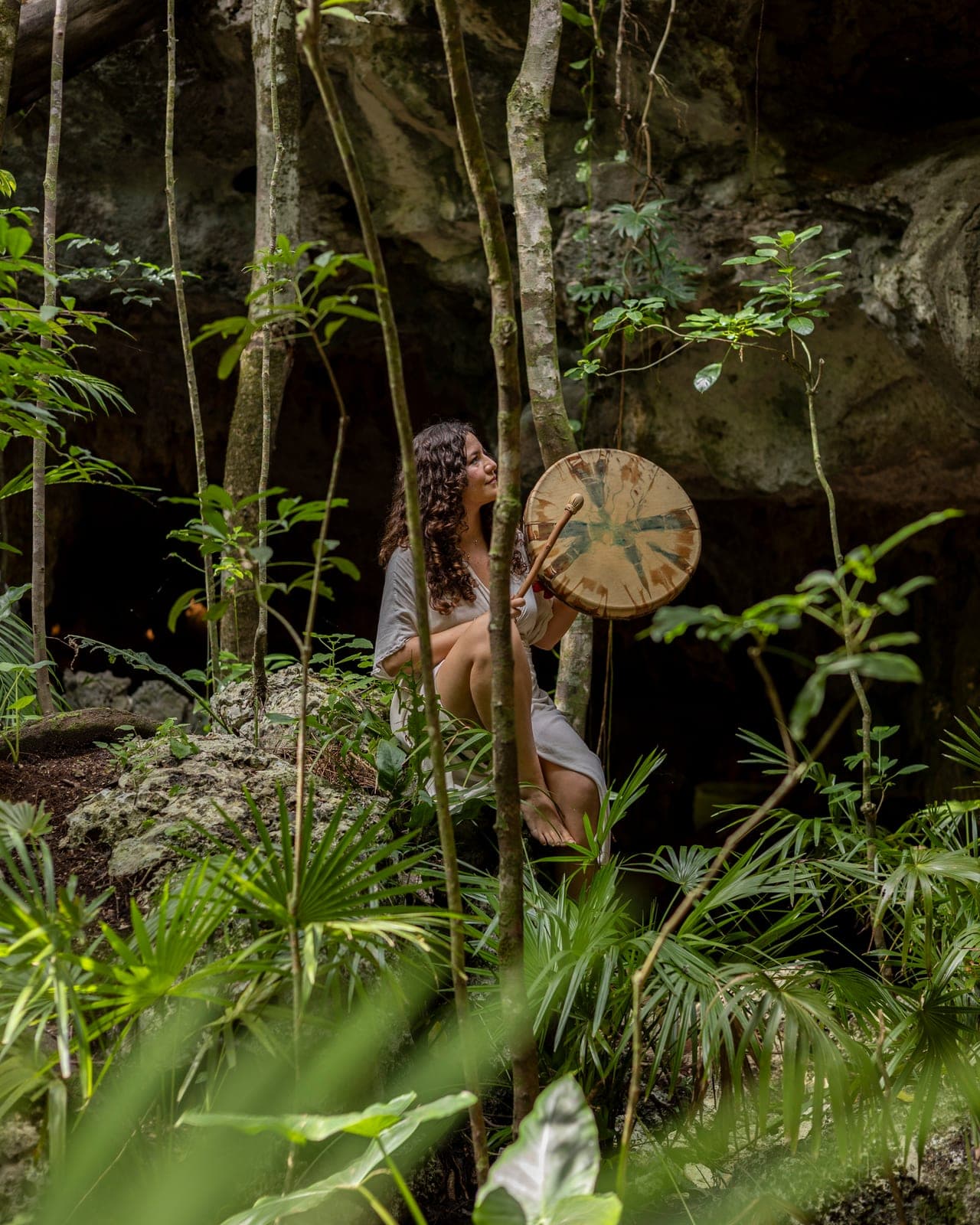 A woman in white seated among palms at the cave mouth, playing a hide frame drum