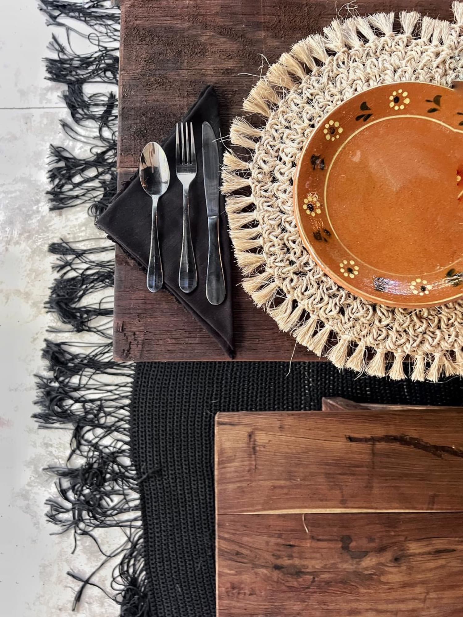 A hand-painted clay plate on a woven charger, black-fringed linen beneath