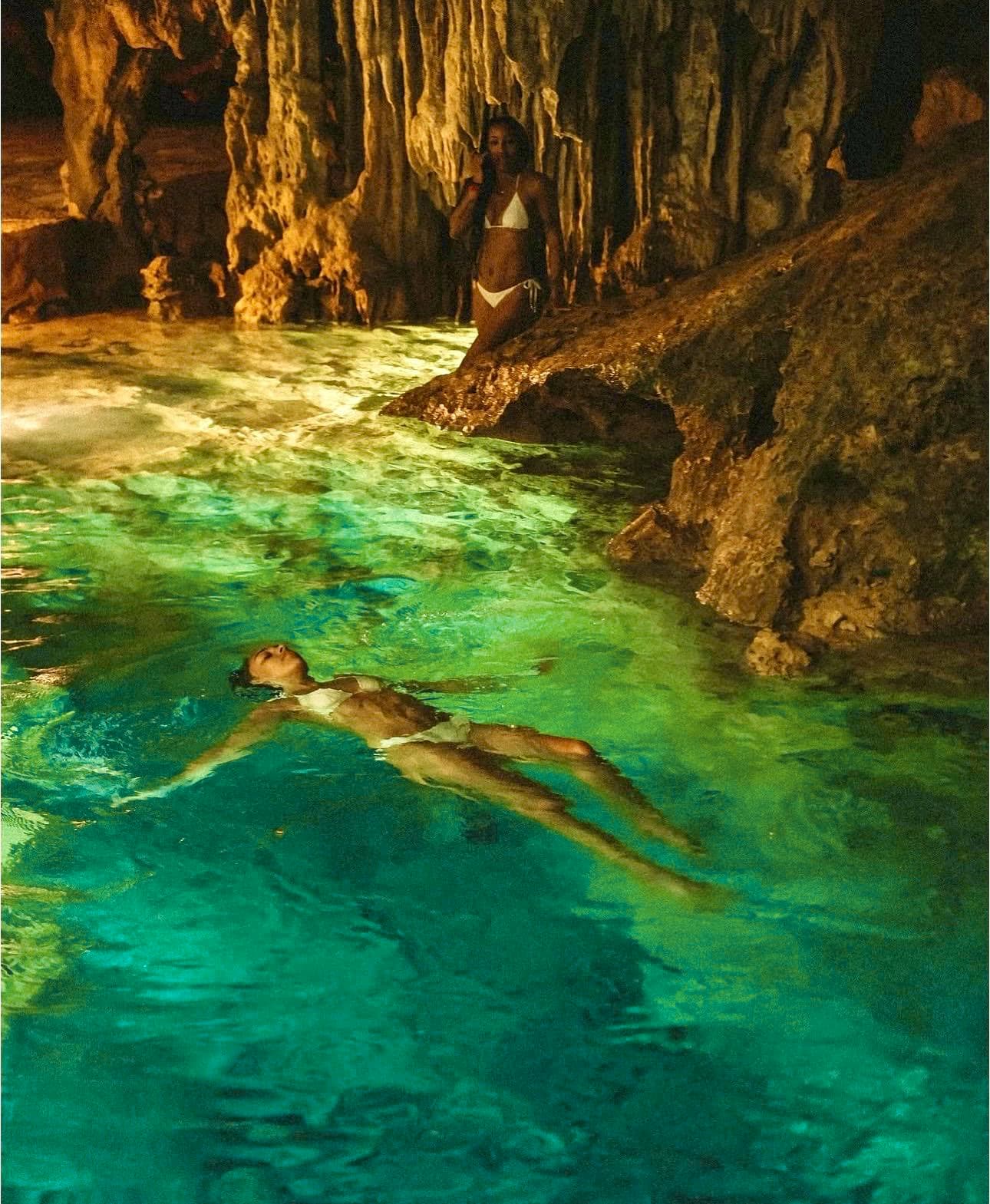 A guest floating on her back across the clear turquoise cenote, the stalactite ceiling above her