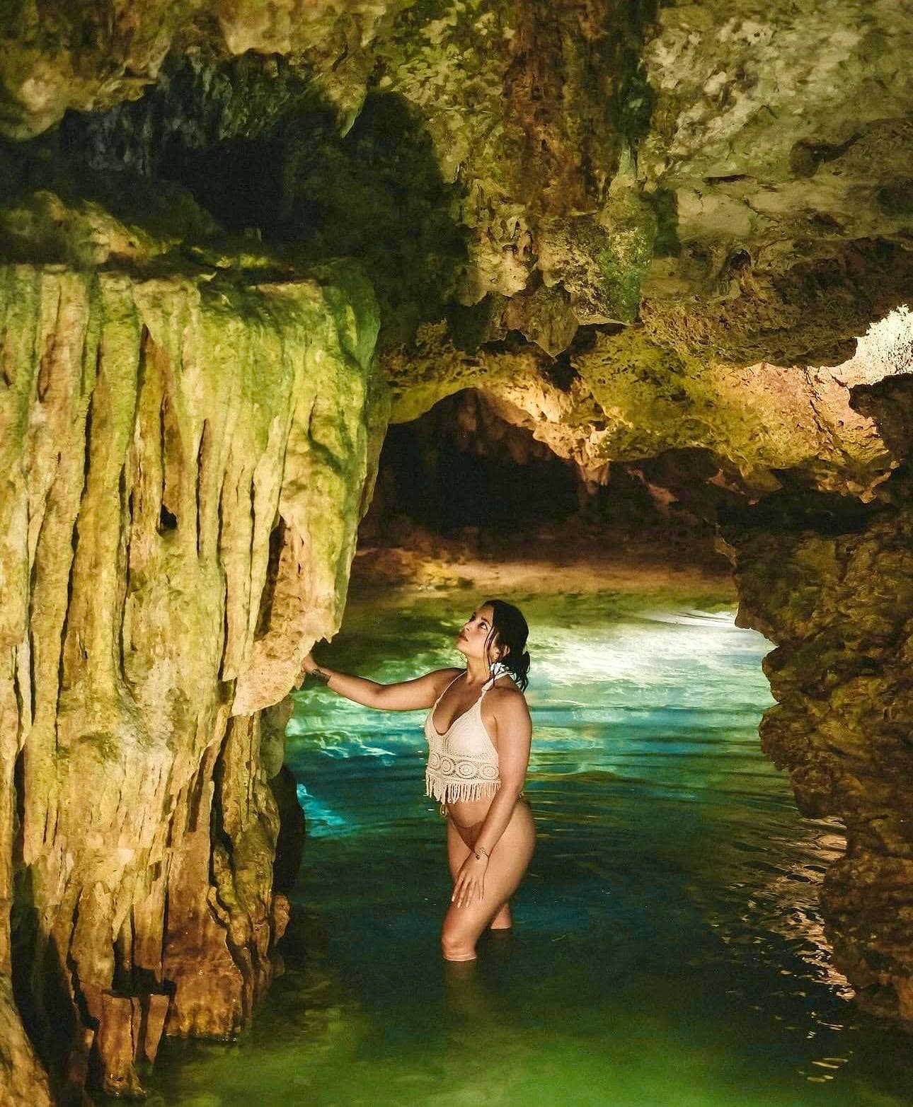A guest standing waist deep in the cenote with a hand on the limestone column, light on the water beyond