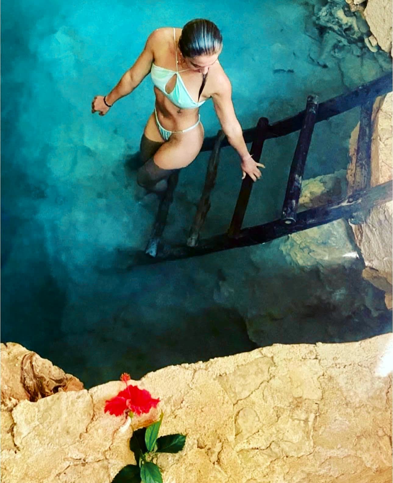 Seen from above, a guest climbing the wooden ladder out of the turquoise cenote, a hibiscus on the stone