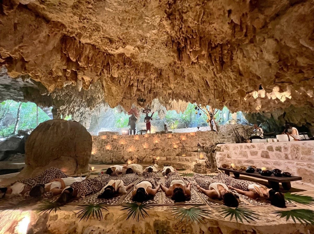 Una ceremonia Quantum en la cueva: huéspedes recostados sobre tapetes bajo el techo de estalactitas, velas a lo largo del muro de piedra y dos Medicine Musicians tocando en la abertura hacia la selva