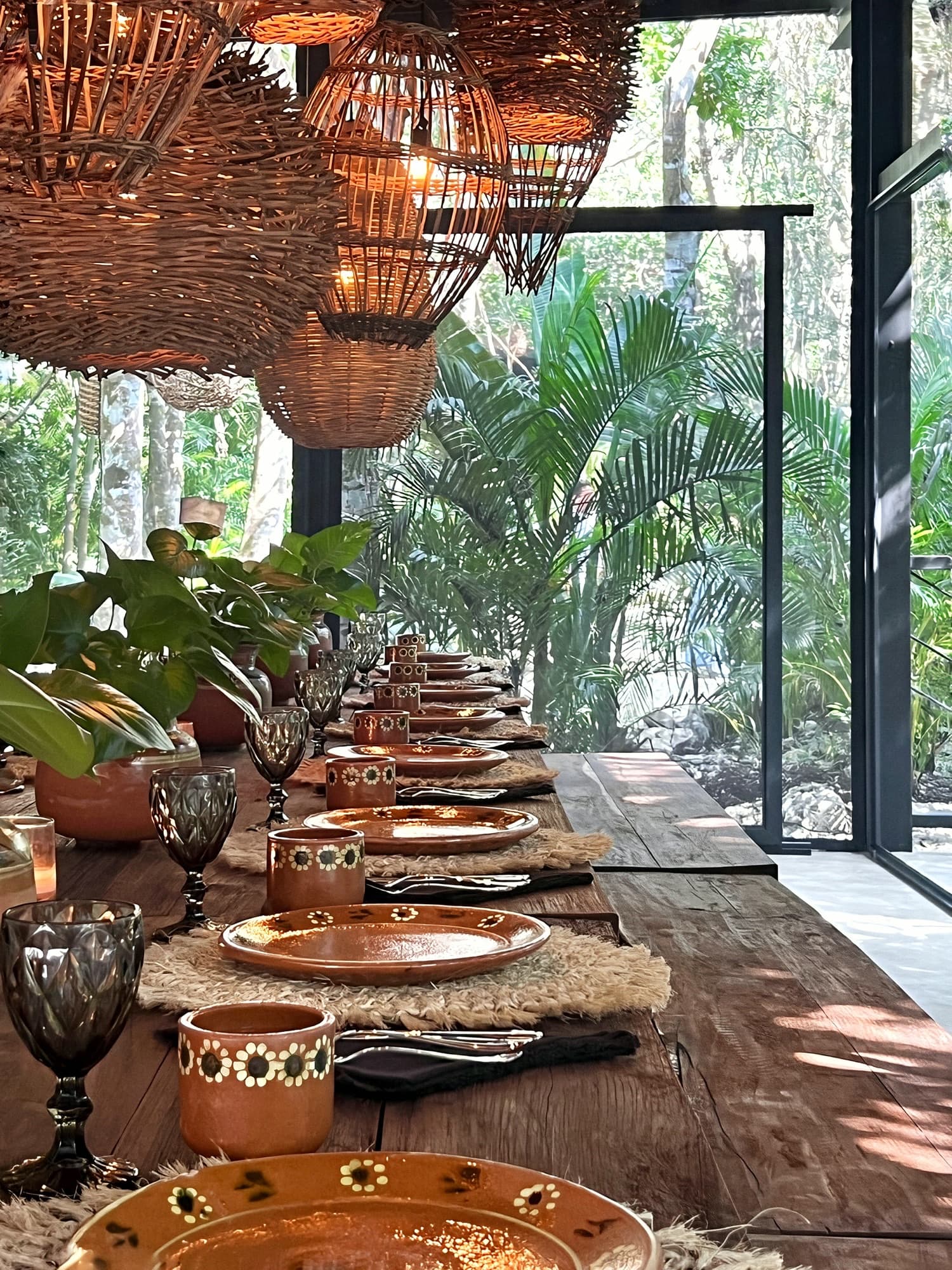 La Mesa laid for dinner: hand-painted clay plates and cups down the long wooden table, woven lanterns overhead and jungle through the glass