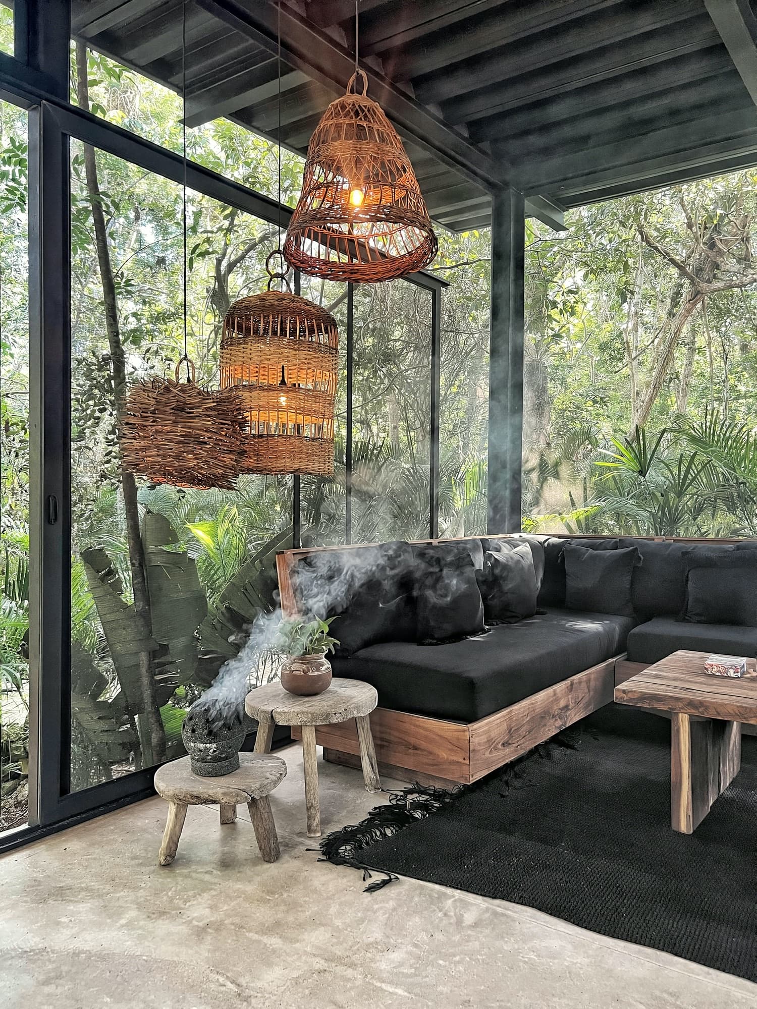 La Recepción: woven lanterns lit above the black sofas, copal smoke rising from the burner, jungle through the glass on every side